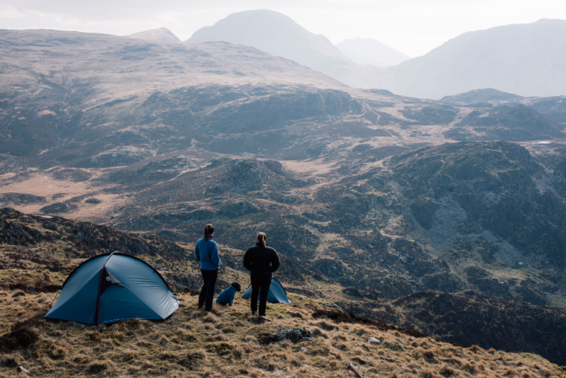 Two people and a dog stand beside blue tents on a grassy mountain ridge, looking out over layered rocky hills and misty peaks under a pale sky.