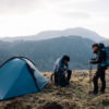 Two hikers beside a blue dome tent on a grassy mountain ridge; one sits adjusting a backpack while the other stands with trekking poles and a large rucksack against a backdrop of layered craggy peaks.