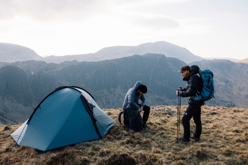 Two hikers beside a blue dome tent on a grassy mountain ridge; one sits adjusting a backpack while the other stands with trekking poles and a large rucksack against a backdrop of layered craggy peaks.