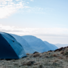 Dark blue camping tent pitched on a windswept grassy hillside with layered blue mountains receding under a pale, cloud-streaked sky.