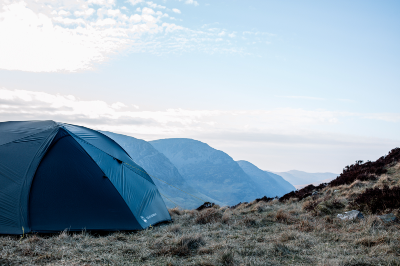 Dark blue camping tent pitched on a windswept grassy hillside with layered blue mountains receding under a pale, cloud-streaked sky.