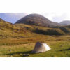 A solitary small grey dome tent pitched on green grass in a wide mountain valley, with rocky slopes and sunlit grassy hills rising behind under a pale blue sky.