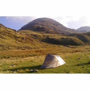 A solitary small grey dome tent pitched on green grass in a wide mountain valley, with rocky slopes and sunlit grassy hills rising behind under a pale blue sky.