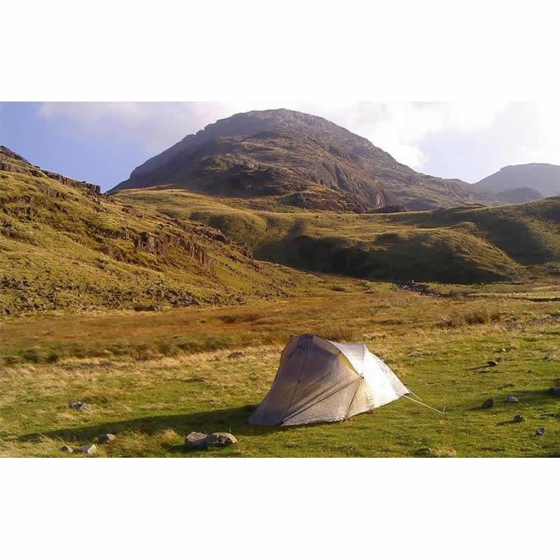 A solitary small grey dome tent pitched on green grass in a wide mountain valley, with rocky slopes and sunlit grassy hills rising behind under a pale blue sky.
