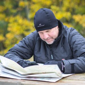 Man wearing a black Extremities beanie, gloves and insulated jacket leans over an open map on a wooden picnic table while checking his phone, with yellow autumn trees blurred in the background.