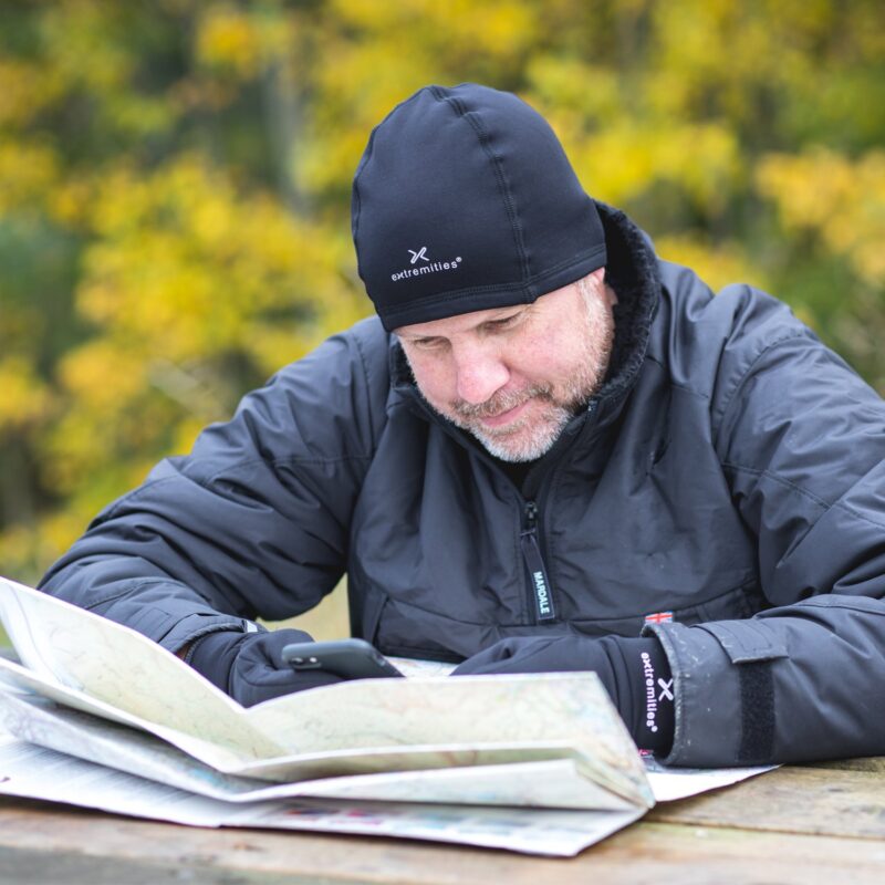 Man wearing a black Extremities beanie, gloves and insulated jacket leans over an open map on a wooden picnic table while checking his phone, with yellow autumn trees blurred in the background.