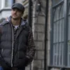 Man wearing a black puffer vest and cap walking casually on a cobblestone street, with historic buildings and large windows in the background.