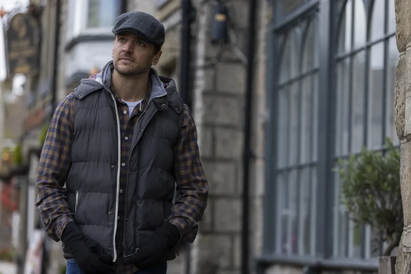 Man wearing a black puffer vest and cap walking casually on a cobblestone street, with historic buildings and large windows in the background.