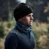 Man wearing a black beanie and a blue scarf, standing outdoors in a natural setting, with trees in the background, showcasing active outdoor apparel suitable for cold weather.