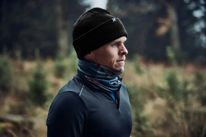 Man wearing a black beanie and a blue scarf, standing outdoors in a natural setting, with trees in the background, showcasing active outdoor apparel suitable for cold weather.