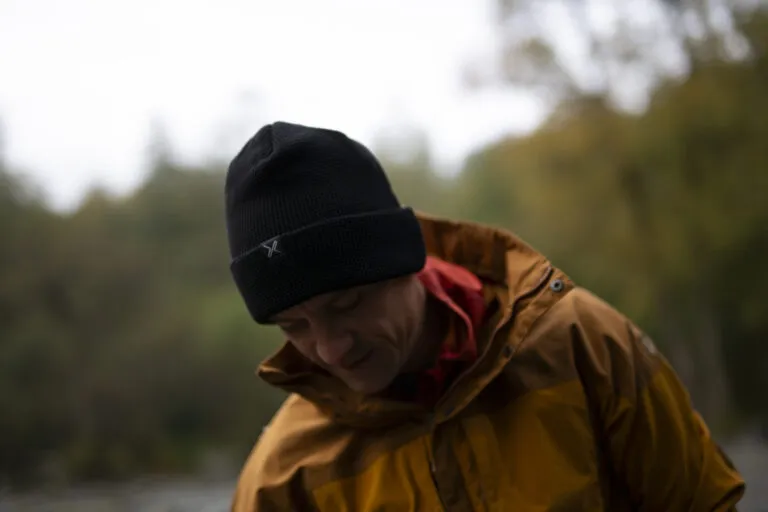 A person wearing a black beanie and a yellow rain jacket, looking down thoughtfully in an outdoor setting with blurred greenery in the background.
