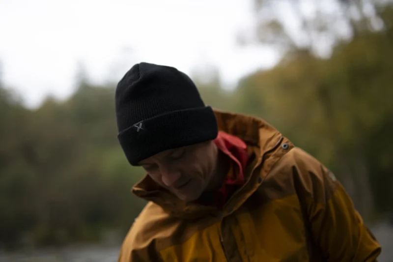 A person wearing a black beanie and a yellow rain jacket, looking down thoughtfully in an outdoor setting with blurred greenery in the background.