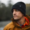 Man wearing a black beanie and orange jacket, looking thoughtfully into the distance in an outdoor setting.