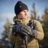 Man wearing a warm jacket and gloves, holding trekking poles, standing outdoors in a forested area with a clear sky.