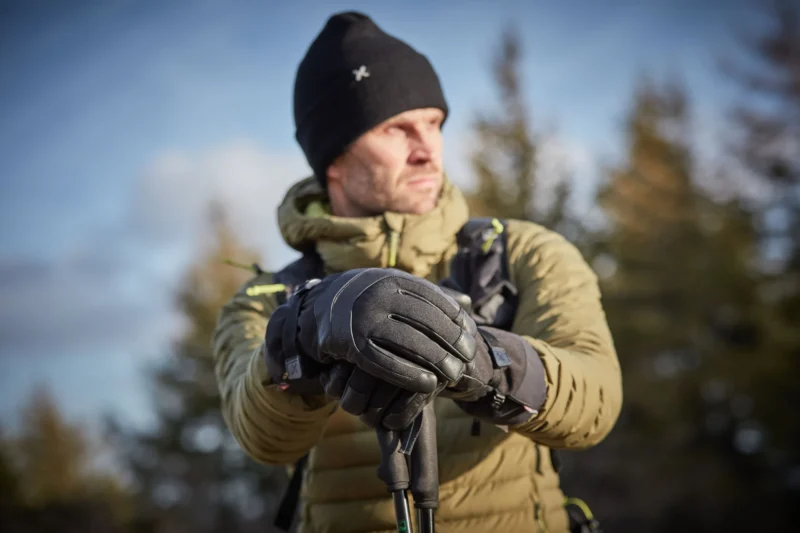 Man wearing a warm jacket and gloves, holding trekking poles, standing outdoors in a forested area with a clear sky.