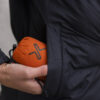 Person holding a compact orange portable speaker while placing it into the pocket of a black jacket, showcasing convenience and style for outdoor activities.