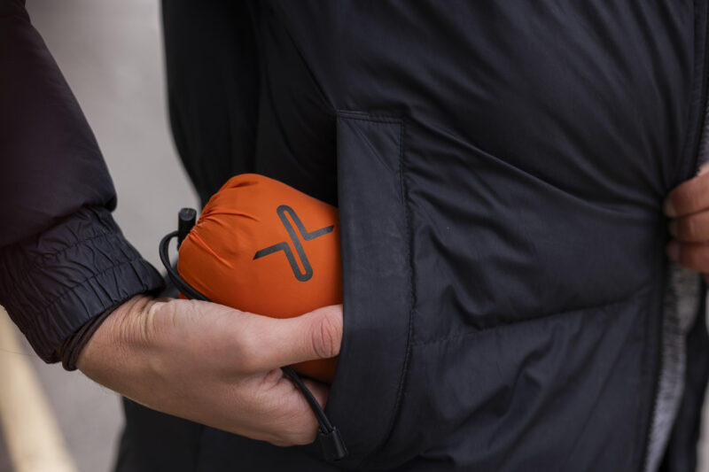 Person holding a compact orange portable speaker while placing it into the pocket of a black jacket, showcasing convenience and style for outdoor activities.