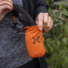 Person holding a compact orange pouch with a black drawstring, designed for easy portability, set against a natural outdoor background.