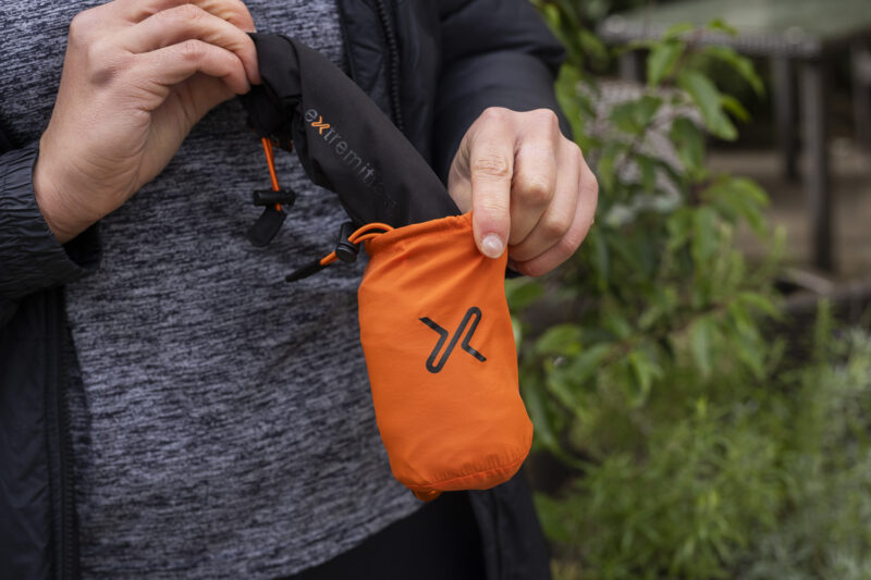 Person holding a compact orange pouch with a black drawstring, designed for easy portability, set against a natural outdoor background.