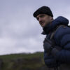Man in a blue puffer jacket and black beanie standing outdoors against a cloudy sky, showcasing a rugged landscape. Ideal for adventure and outdoor gear content.