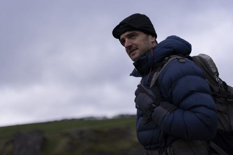 Man in a blue puffer jacket and black beanie standing outdoors against a cloudy sky, showcasing a rugged landscape. Ideal for adventure and outdoor gear content.