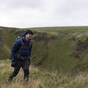A hiker in a blue puffer jacket and black hat walks through grassy terrain near a cliff, surrounded by rolling green hills under a cloudy sky.