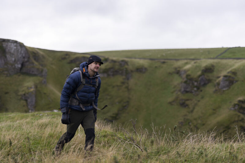 A hiker in a blue puffer jacket and black hat walks through grassy terrain near a cliff, surrounded by rolling green hills under a cloudy sky.