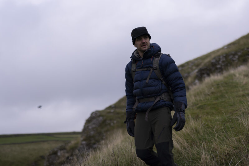 Man hiking on a grassy hillside wearing a blue puffer jacket, gloves, and a hat, with a cloudy sky in the background.